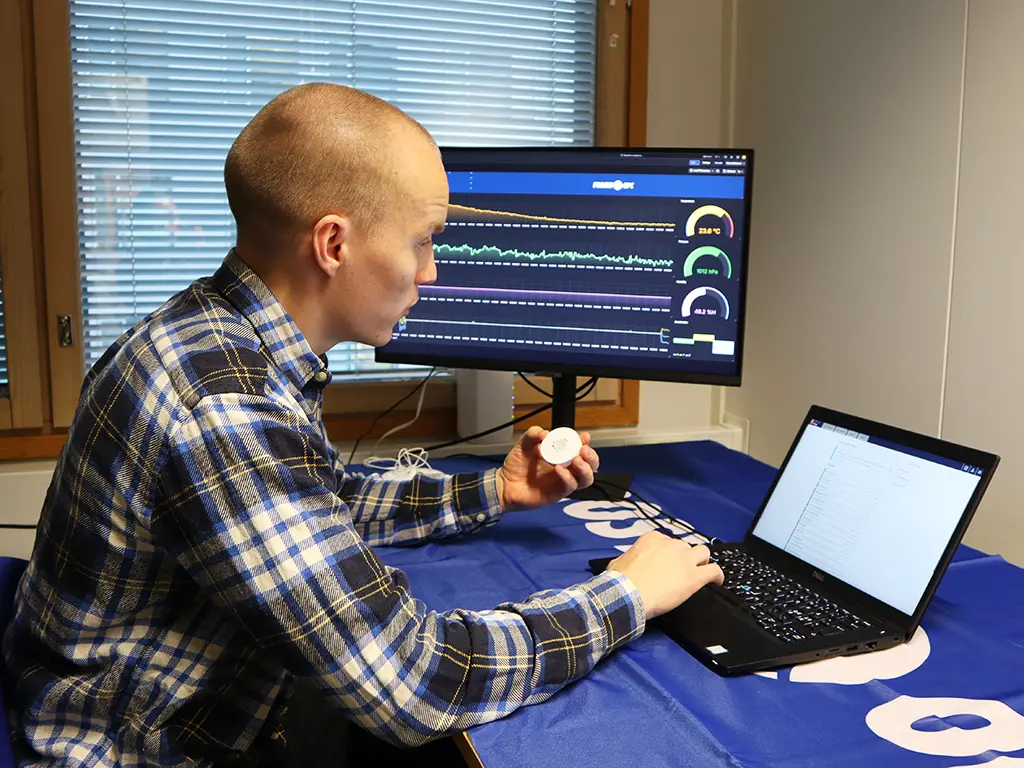 Man sitting at a computer examining a Ruuvi sensor while viewing Grafana charts and OPC UA Forge on the screen.