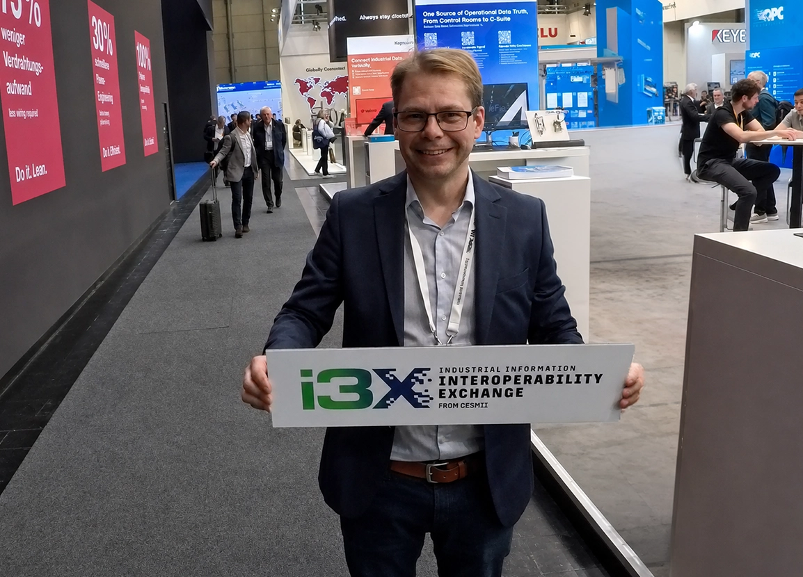 Man in a navy blazer holding a white sign with 'i3X INTEROPERABILITY EXCHANGE' at a trade show.